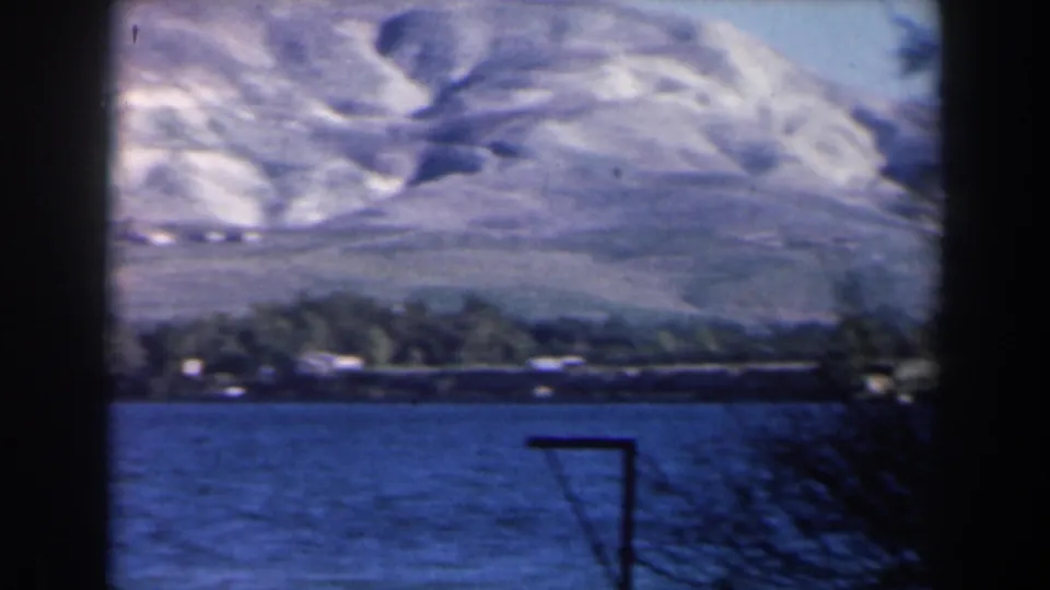 1960 Israel home-movie frame showing blue river water, a low settlement or shoreline buildings, and pale mountains in the background.