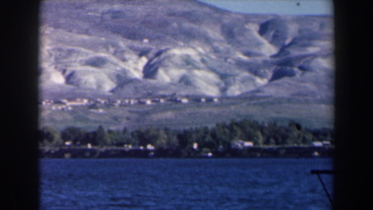 1960 8mm view in Israel across blue lake water toward trees, distant buildings, and eroded hills.