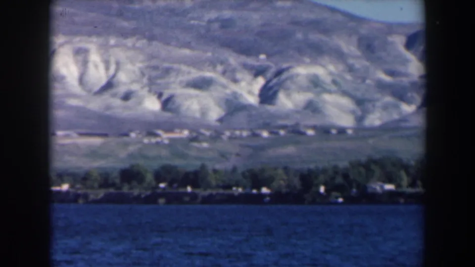 1960 8mm Israel landscape with lake foreground, low shoreline structures, trees, and pale ridged hills.