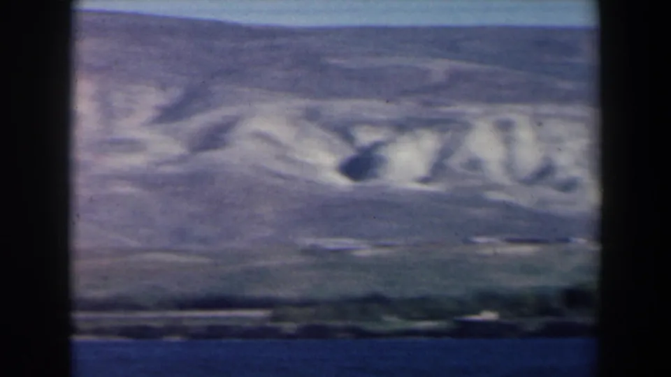 Soft 1960 8mm frame of an Israel lakeside landscape with water, low shore, and pale eroded hills.