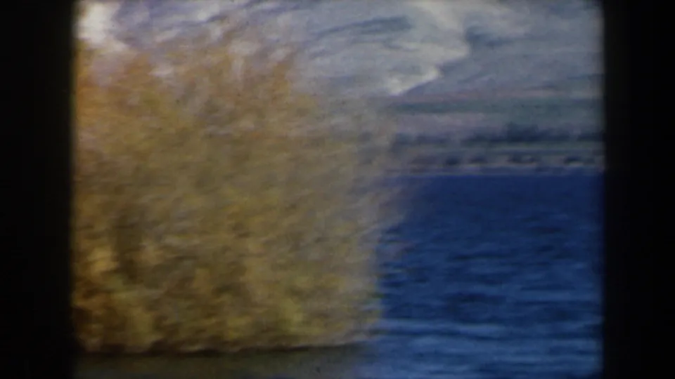 1960 8mm Israel landscape with blue lake water, shoreline trees, and pale mountain ridges in the background.