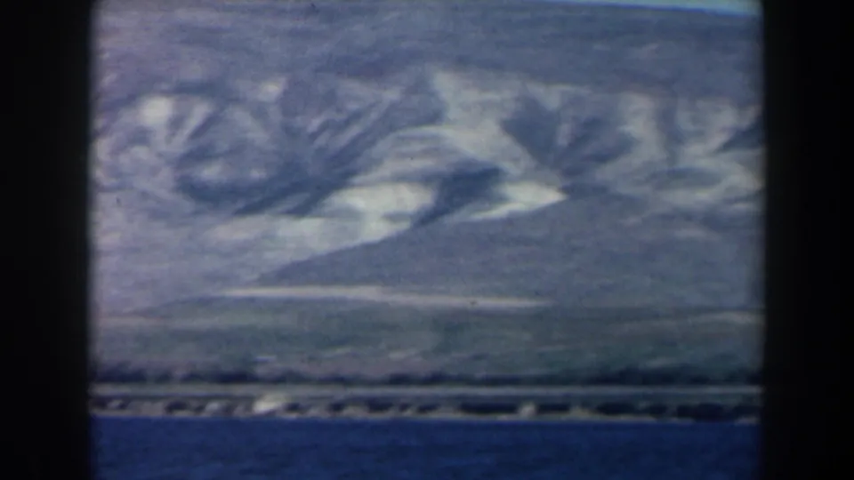 1960 8mm Israel frame showing a broad lake foreground with pale mountain ridges and a distant shoreline across the water.