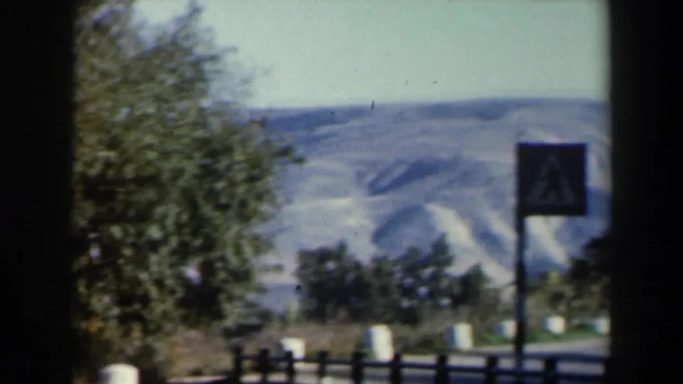 1960 8mm Israel overlook frame with a fence, roadside-style sign, trees, and pale hills beyond the lake area.