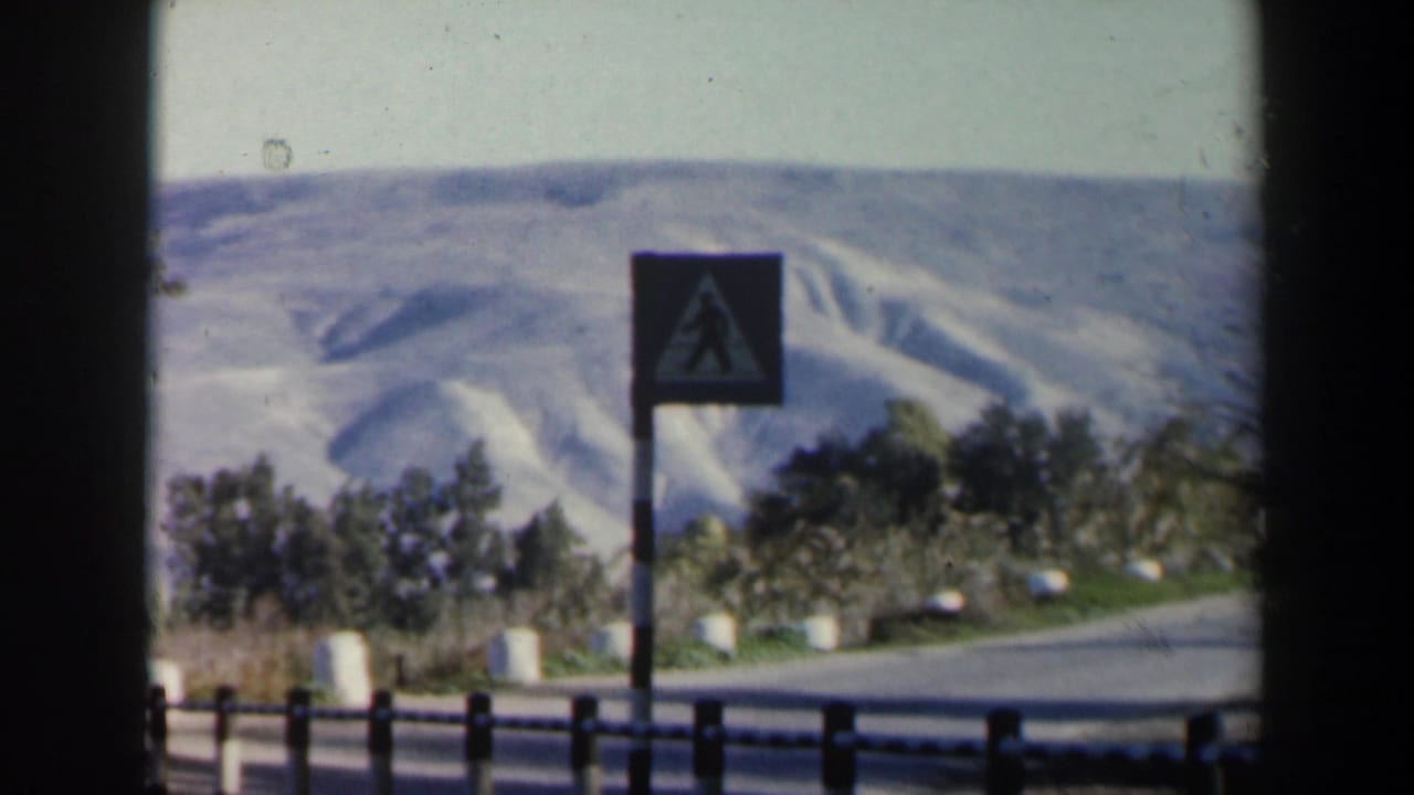 1960 Israel 8mm frame showing a pedestrian crossing sign near a winding road, white barriers, trees, and pale mountain slopes.