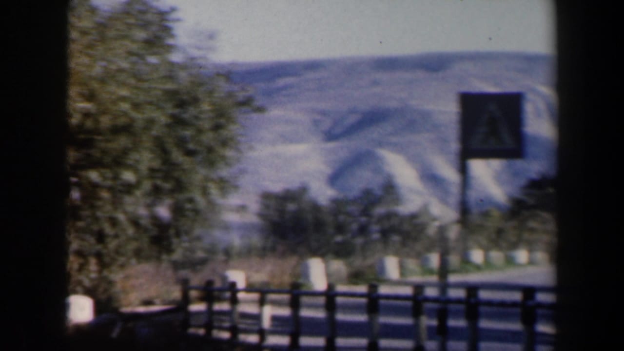 Roadside sign and fence seen from a vehicle with hills behind trees.