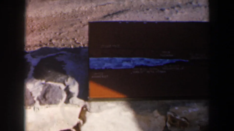1960 Israel 8mm frame showing a brown tackle box with a blue stripe on grey rocks, partly obscured by dark frame edges.