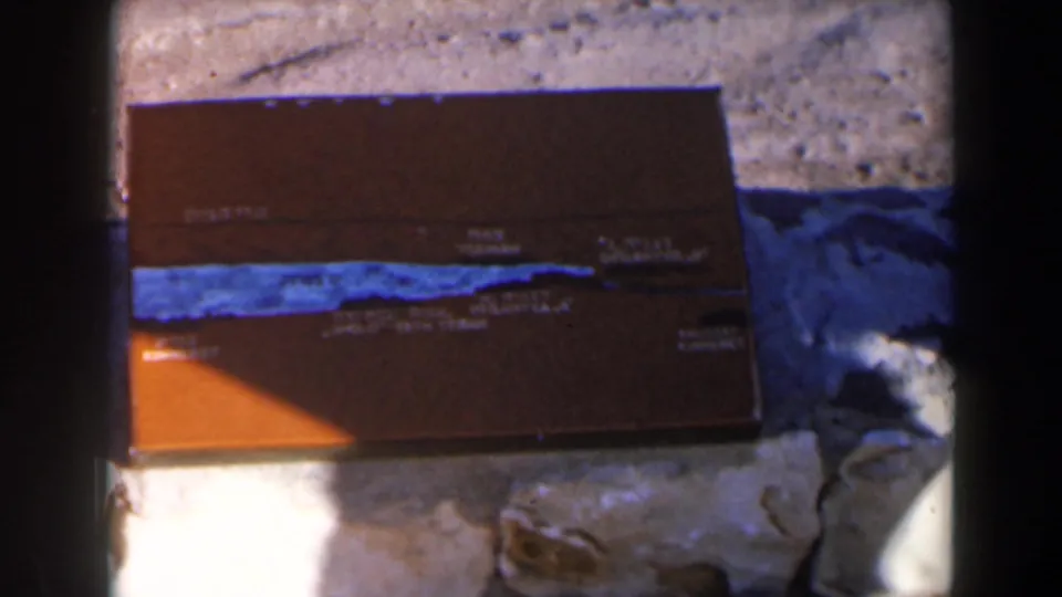 1960 Israel 8mm frame with a closer view of the brown tackle box, blue stripe, faded text, and surrounding grey rocks.