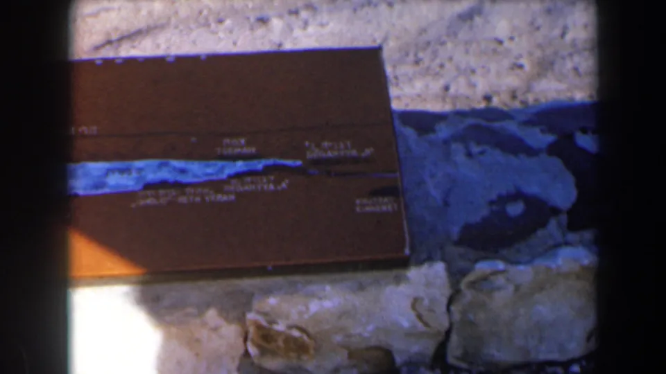1960 Israel 8mm frame showing the brown tackle box on rocks with a blue stripe, faded markings, and dark edge framing.