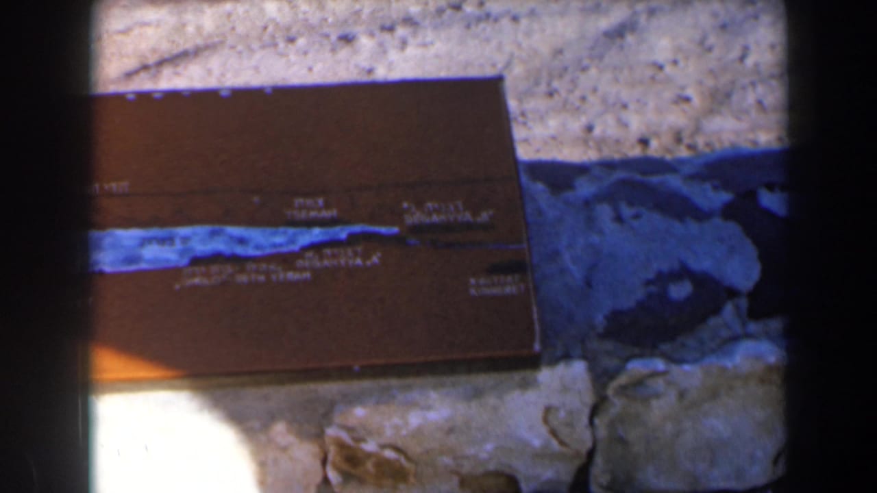Brown bilingual plaque on stone and gravel, partly shadowed with dark film edges.