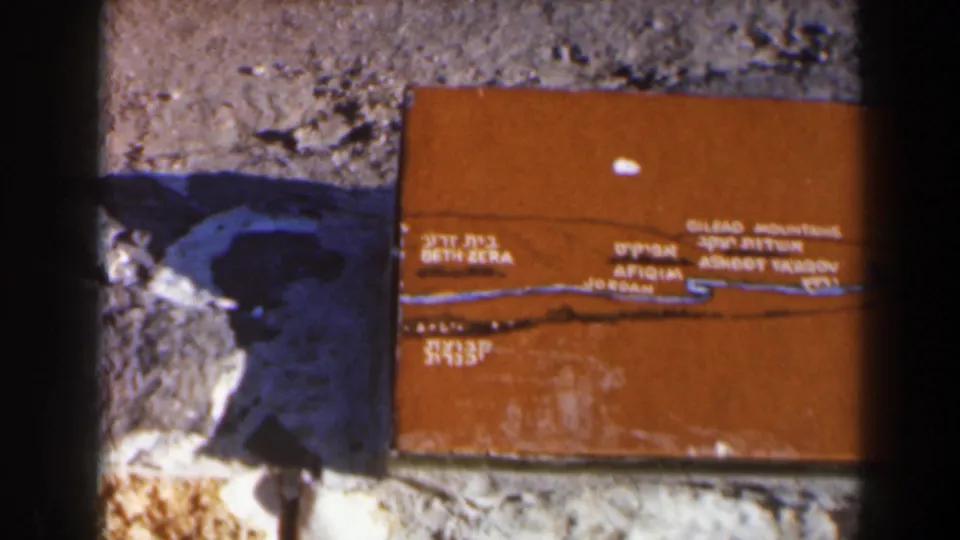 1960 Israel home-movie frame showing a closer view of the plaque text and blue route line beside rough stones.