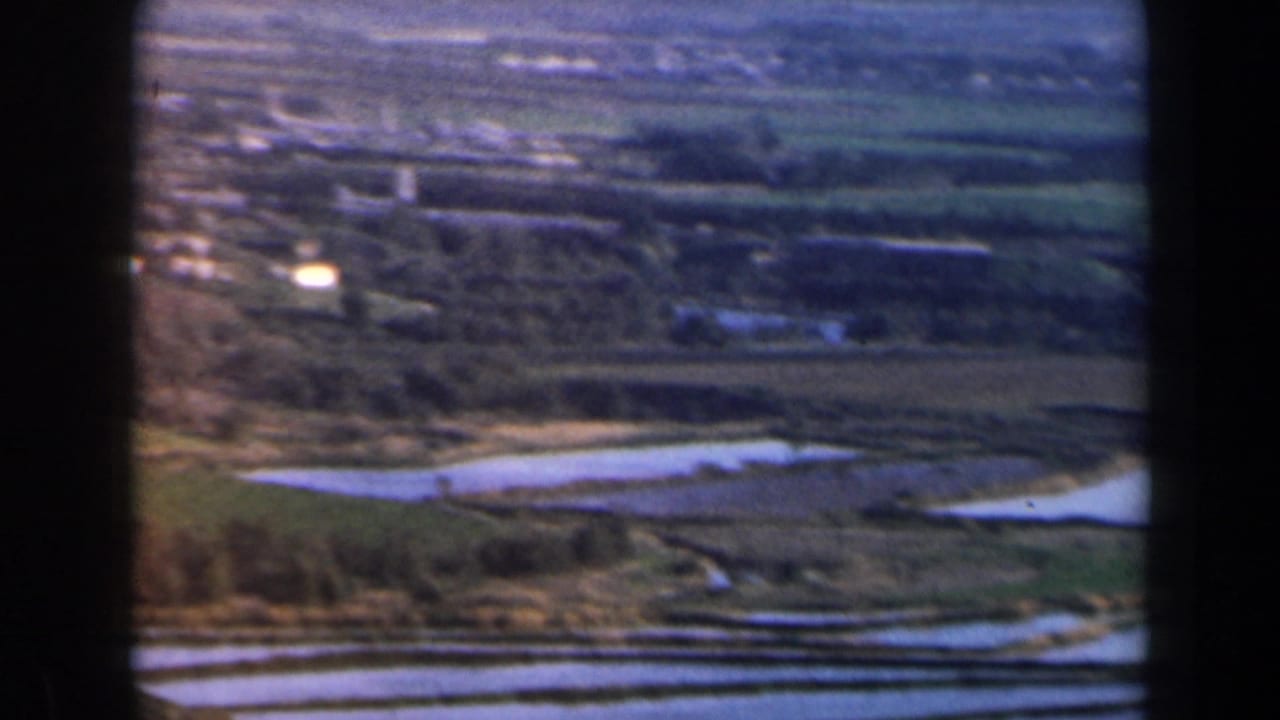 Elevated view of fields, water channels, trees, and distant buildings bordered by dark film edges.