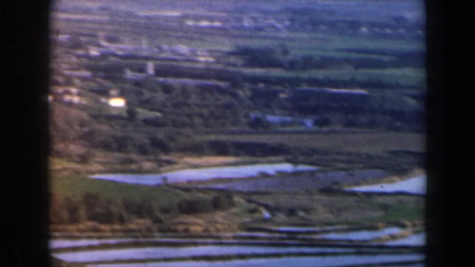 1960 Israel home-movie frame showing a town and rural fields from above, with reflective water channels and dark 8mm frame borders.