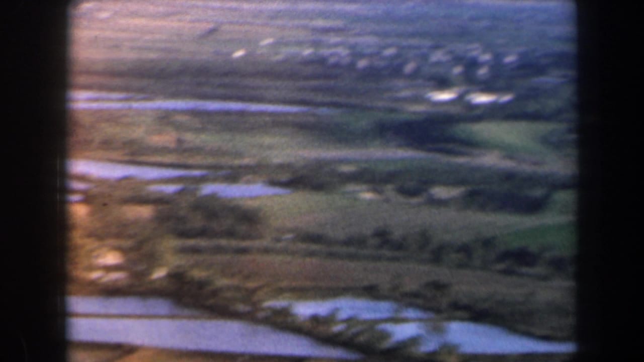 Rolling fields and bright water patches with a distant settlement under soft light.