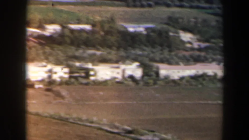 1960 Israel frame showing white low buildings, cultivated fields, trees, and reflective water from an elevated home-movie viewpoint.