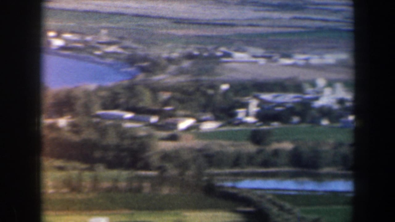 Blurred aerial view of fields, water, trees, and scattered low buildings.