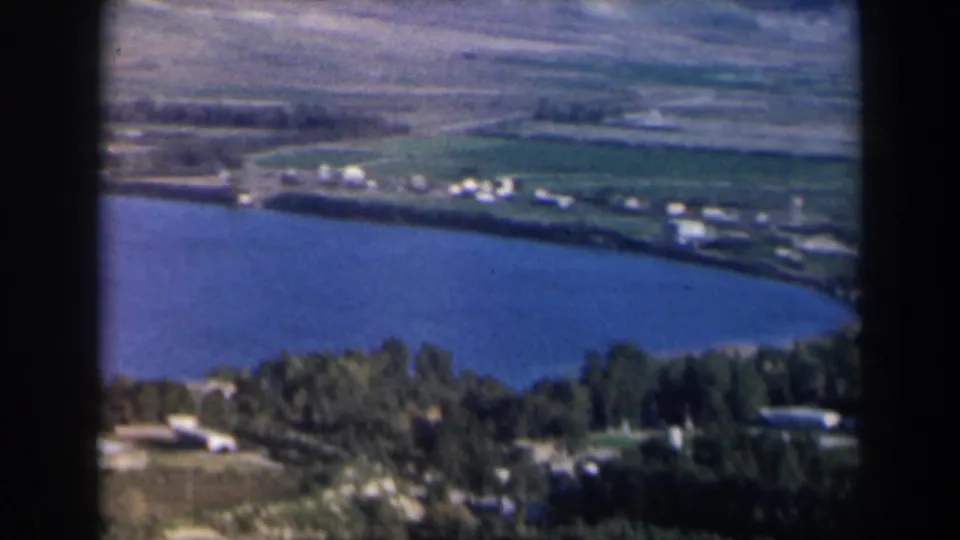 1960 Israel frame showing a broad blue lake bordered by fields, trees, and distant white buildings.
