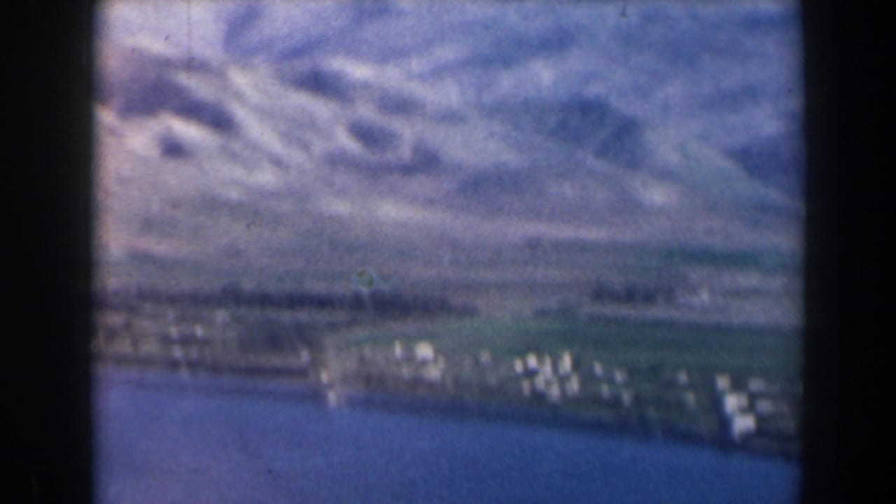 Soft aerial view of blue water, green shoreline fields, white buildings, and pale hills beyond.