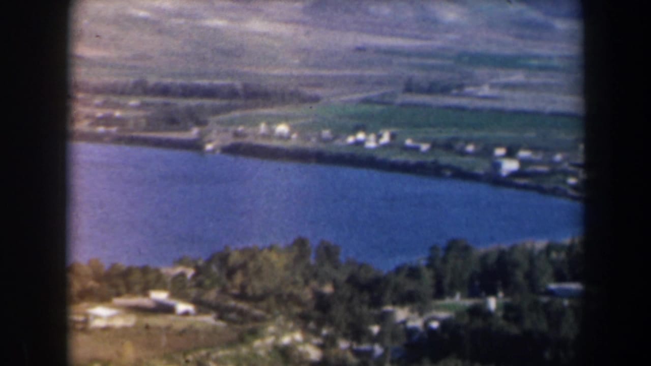 Aerial view of a broad blue lake bordered by trees, fields, small buildings, and dry hills.