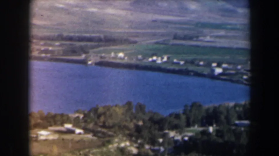 1960 8mm aerial frame from Israel with blue water, a tree-lined shore, cultivated land, scattered buildings, and arid hills behind.