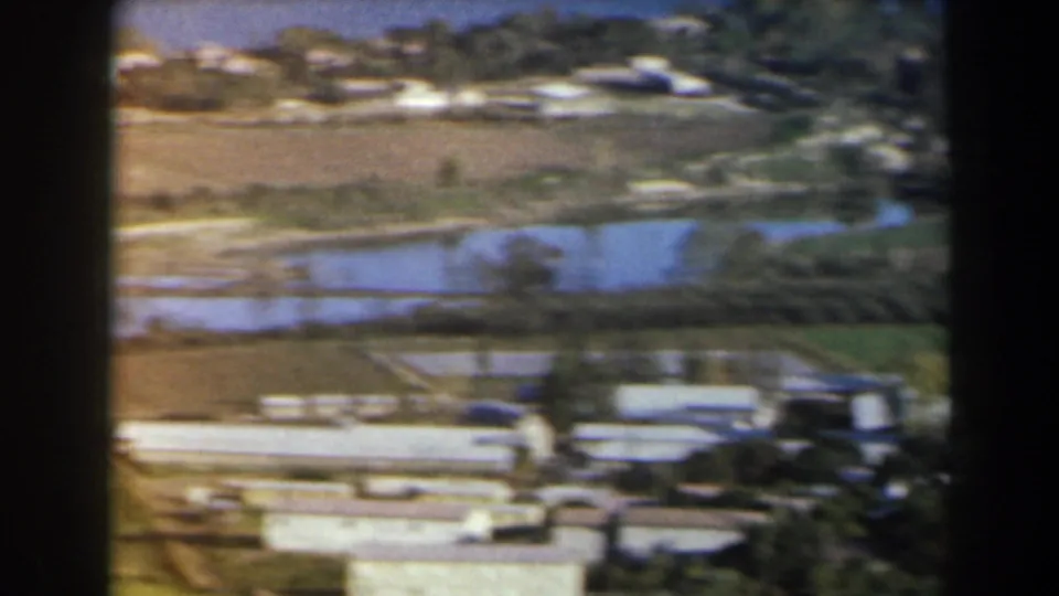 1960 Israeli home-movie frame showing rural fields, reflective water channels or ponds, trees, low buildings, and shoreline in soft focus.