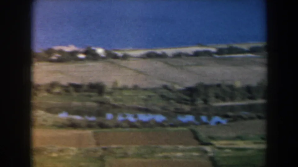 1960 8mm aerial frame in Israel with patterned fields, narrow reflective water features, a pale shoreline, and blue water beyond.