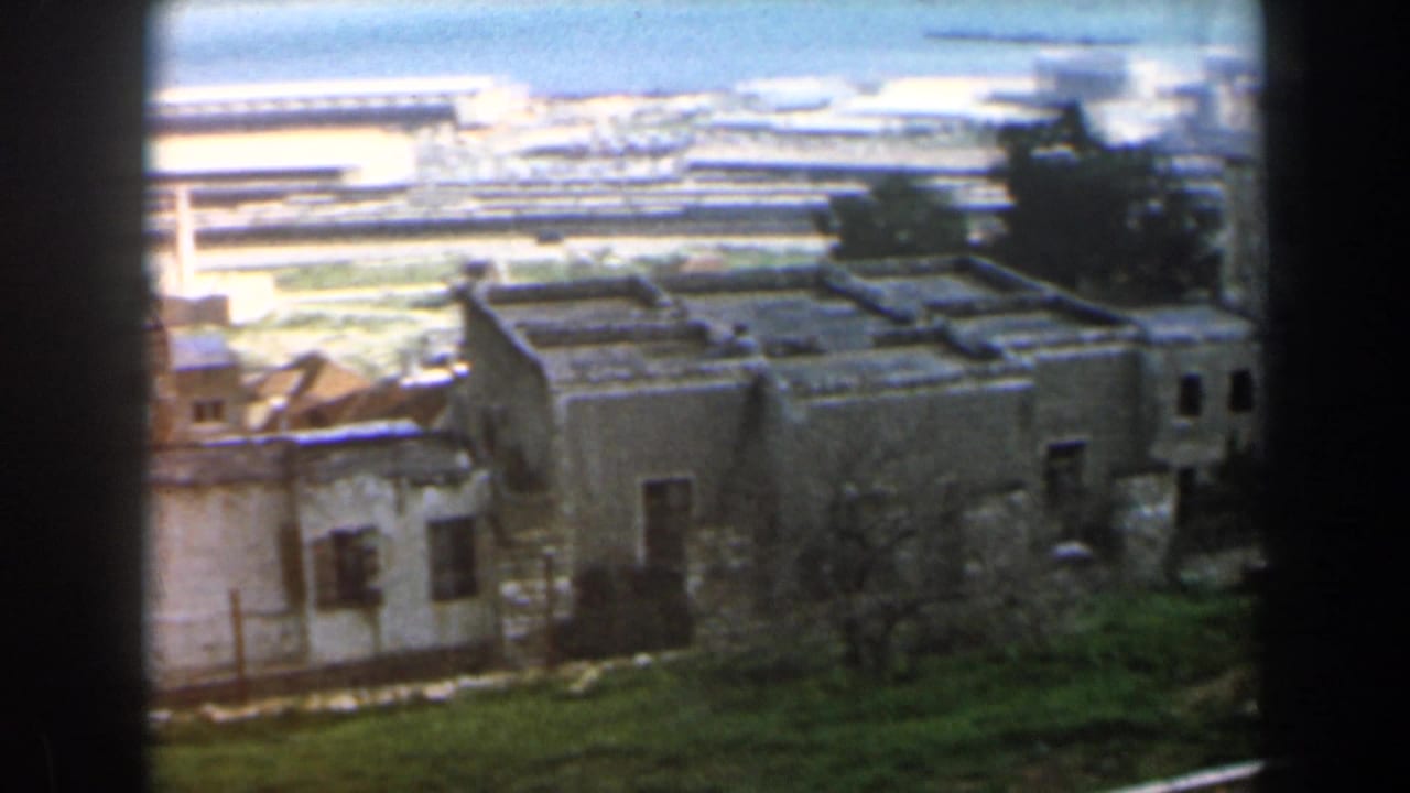 Low stone houses and a taller building seen from above near a distant waterfront.