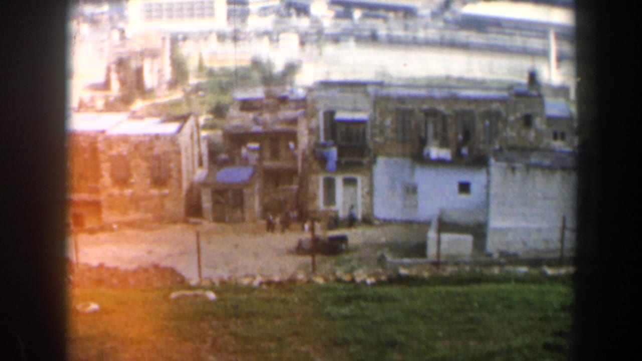 1960 8mm view in Israel showing tan low-rise buildings, open ground, and harbor water in the distance.