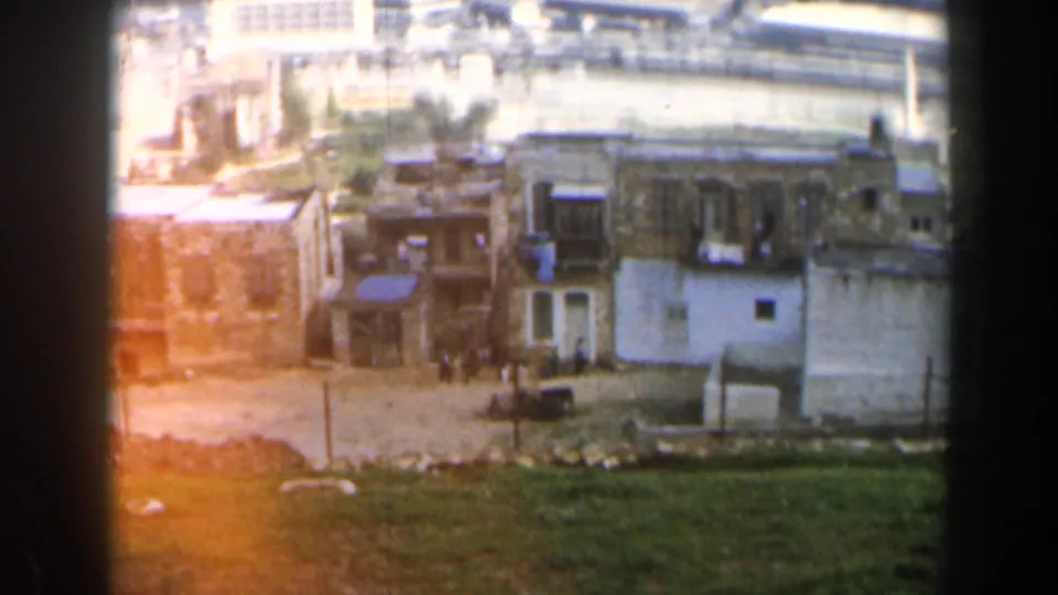 1960 8mm view in Israel showing tan low-rise buildings, open ground, and harbor water in the distance.