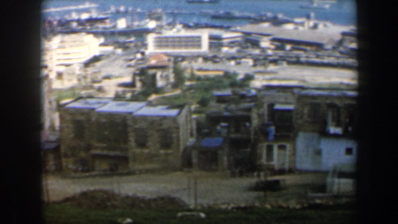 Panoramic view of low buildings, roads, harbor structures, and sea.