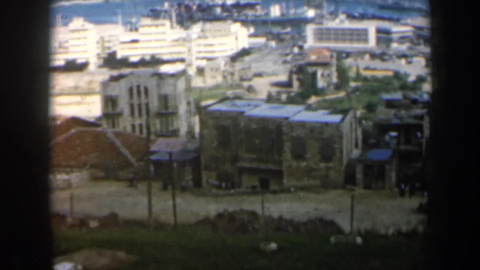 1960 8mm frame from Israel showing stone buildings, a road, and bright harbor-side structures beyond.