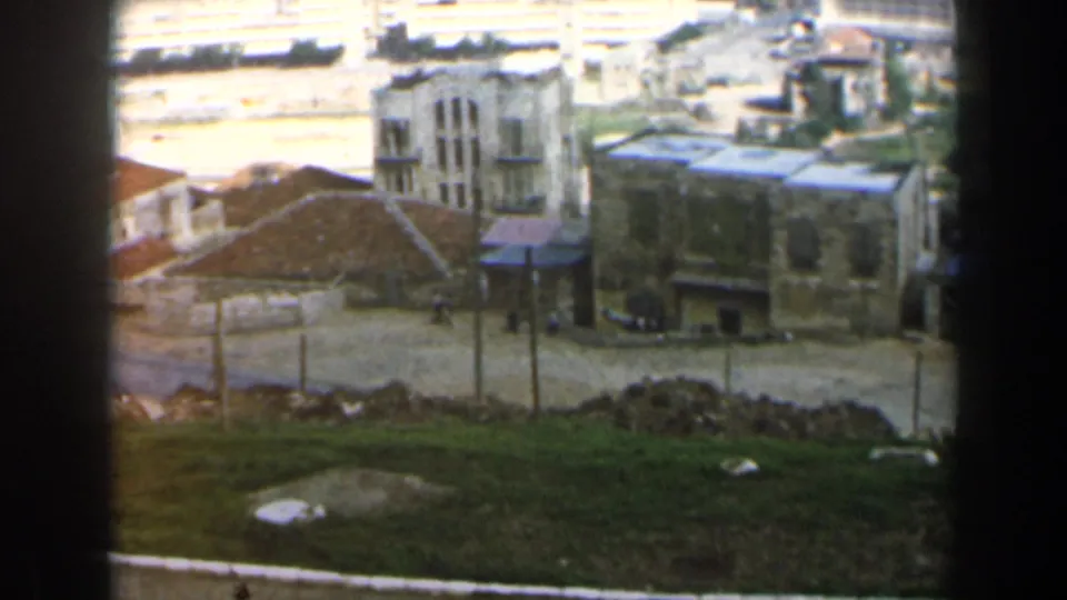 End of the 1960 Israel pan with tan buildings, small distant figures, open ground, and harbor structures behind.