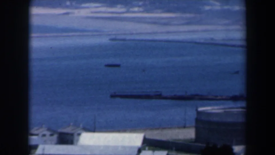 1960 Israel home-movie frame of open coastal water, low foreground roofs, and long barges or cargo vessels offshore.