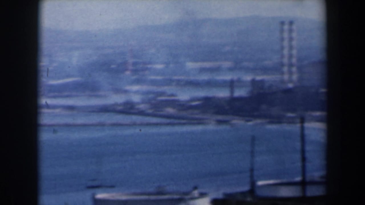 Industrial shoreline with water, storage tanks, and tall smokestacks in haze.