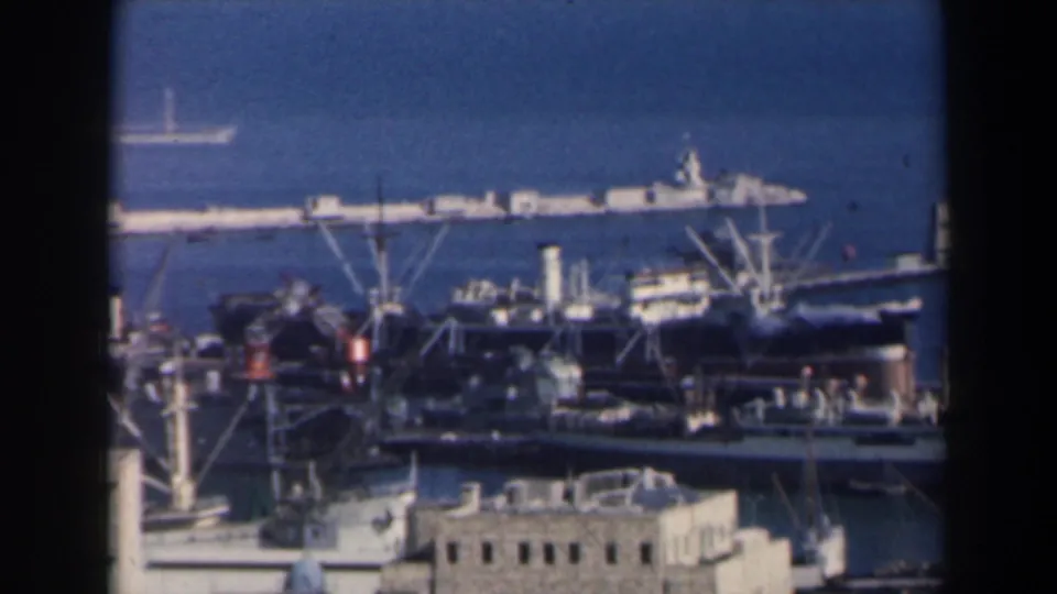 1960 Israel 8mm view of dockside cranes, moored ships, low buildings and a breakwater across the harbor.