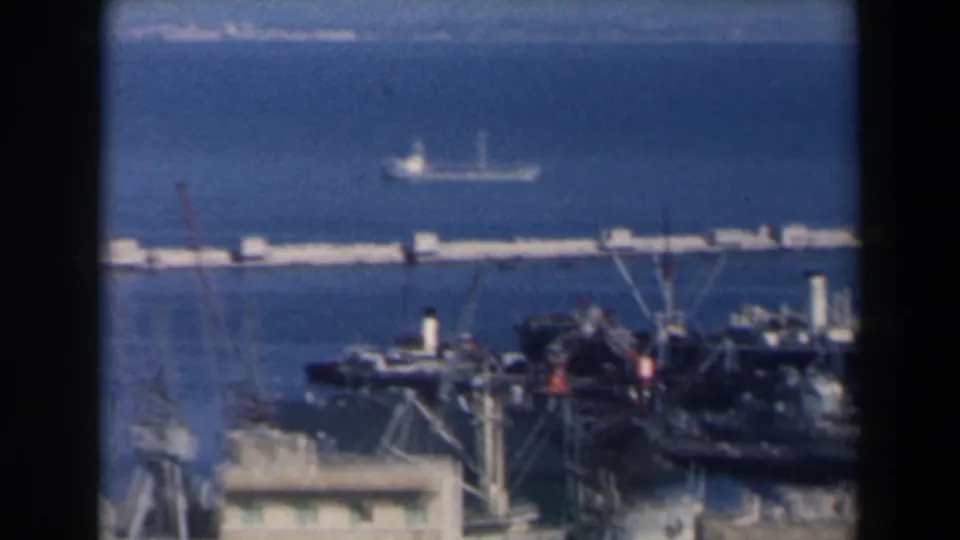 Elevated 1960 Israel harbor frame with cranes, docked vessels, low port buildings and open blue water beyond.