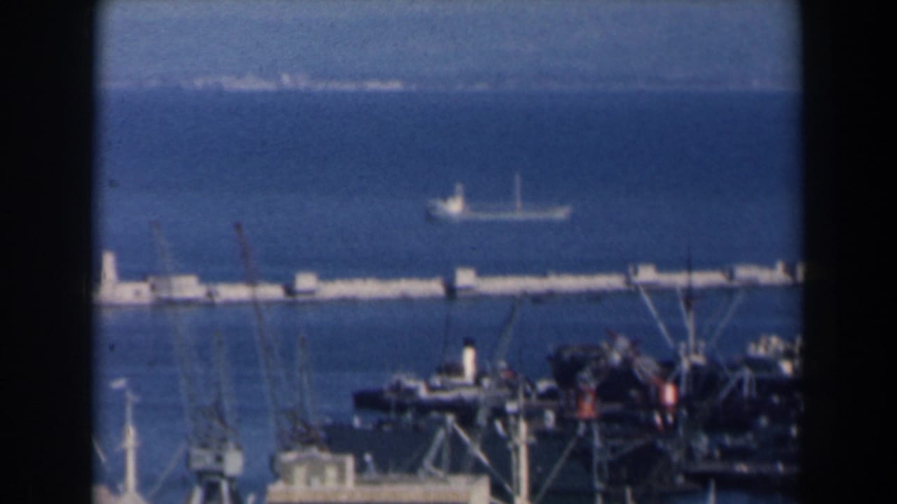 1960 Israel home-movie frame showing a harbor from above with dark foreground edges, cranes, ships, a breakwater, and open sea.