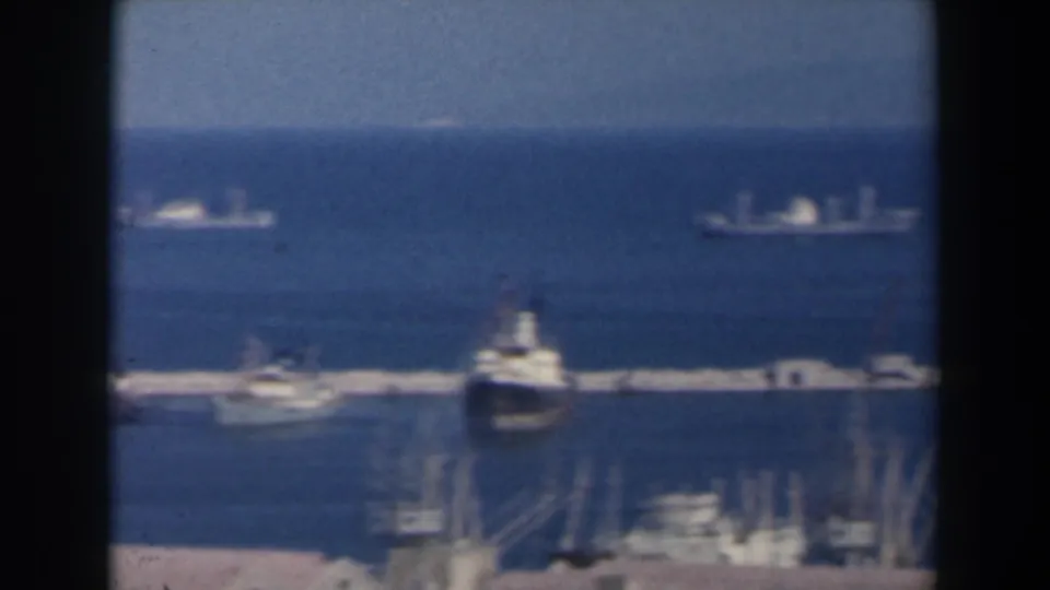 1960 Israel home-movie frame with distant ships on the blue sea, a breakwater, harbor cranes, and soft foreground waterfront detail.