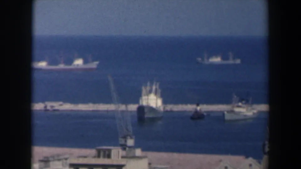 1960 Israel harbor frame showing a breakwater, several distant vessels, small boats in the basin, cranes, and deep blue sea.