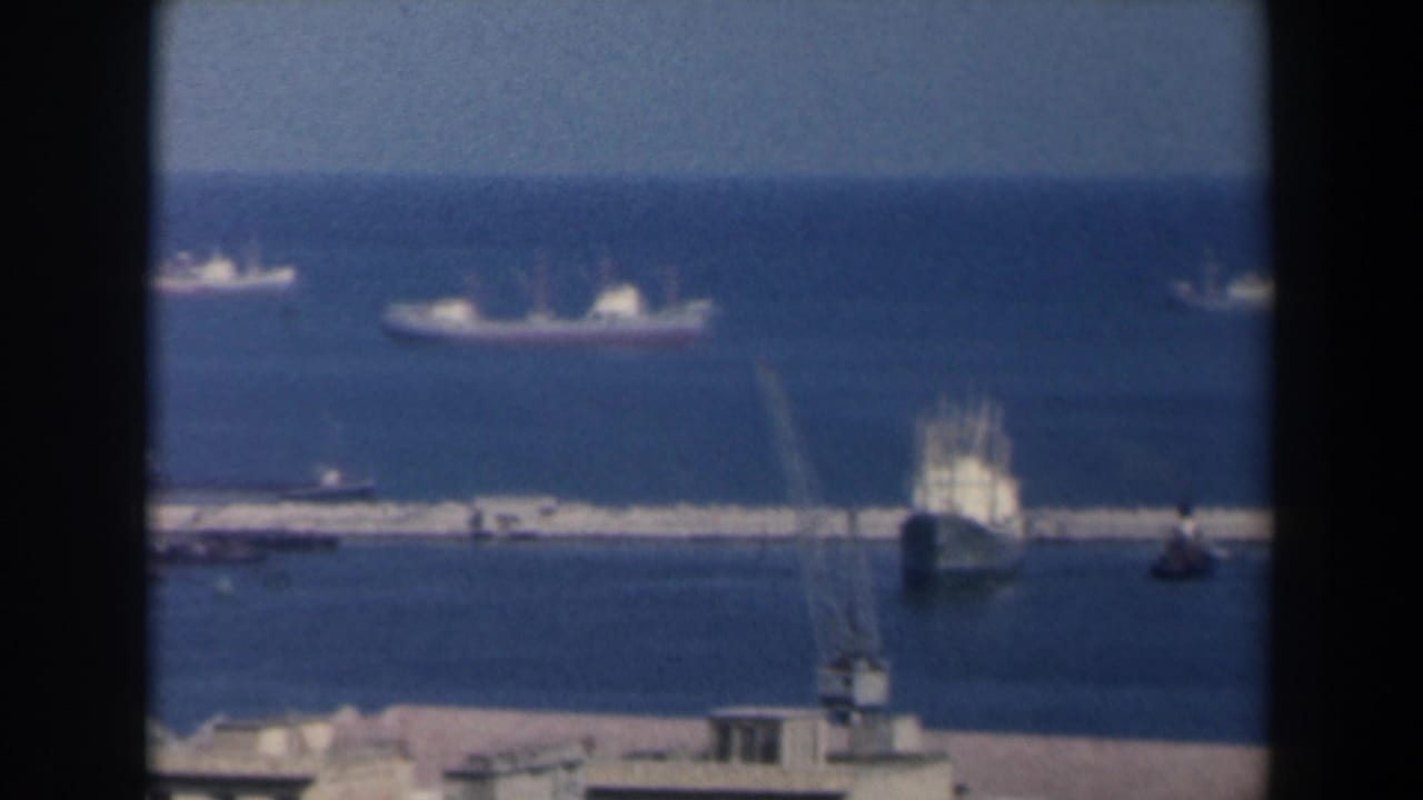 1960 Israel 8mm harbor view with distant ships, a breakwater or pier, and waterfront structures seen from above.
