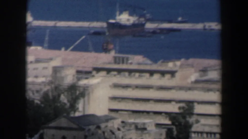 Elevated 1960 Israel harbor view with a vessel on blue water beyond layered rooftops and port structures.