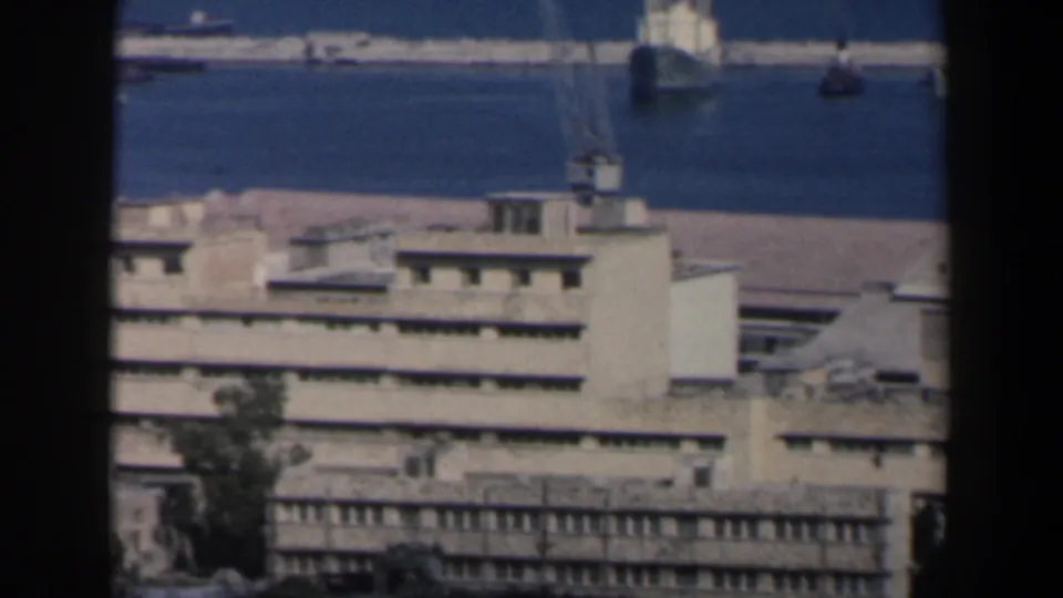 1960 Israel harbor scene with a port building and crane in front of blue water and a small distant boat.