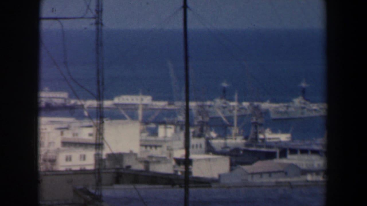 Distant harbor with docked ships, cranes, buildings, and blue sea beyond dark foreground poles.