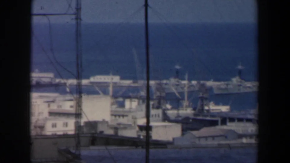 1960 Israel 8mm frame showing a distant harbor with ships, cranes, waterfront buildings, and the sea horizon behind foreground poles.