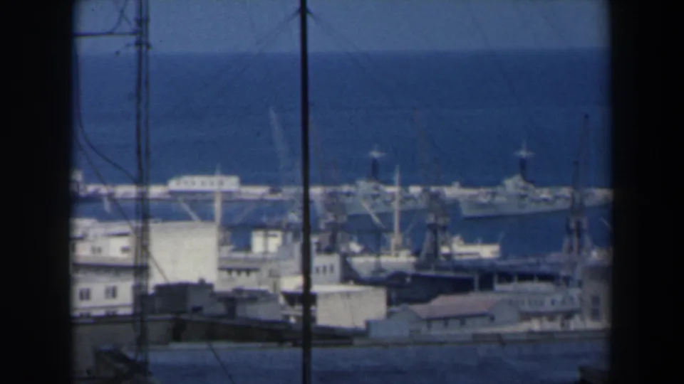 1960 Israel 8mm frame of a wide port scene, with docked ships, cranes, low industrial structures, and blue water beyond.