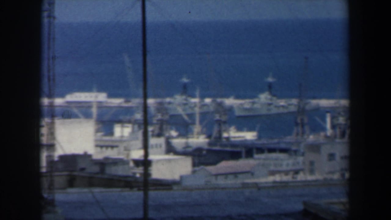 White harbor buildings and docked ships seen through dark window-like edges beside blue sea.