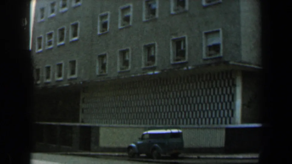 1960 Israel 8mm frame showing a multi-story building exterior, shuttered ground level and a vintage blue car at the curb.