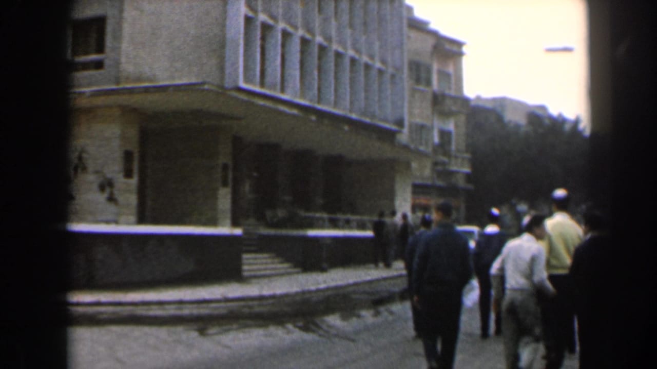 1960 Israel street view with people walking beside a light multi-story building and shaded entrance.