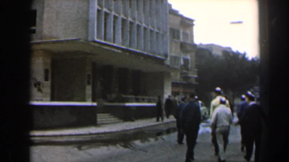 Pedestrians in suits and light shirts move along a 1960 Israel street next to a gridded facade.