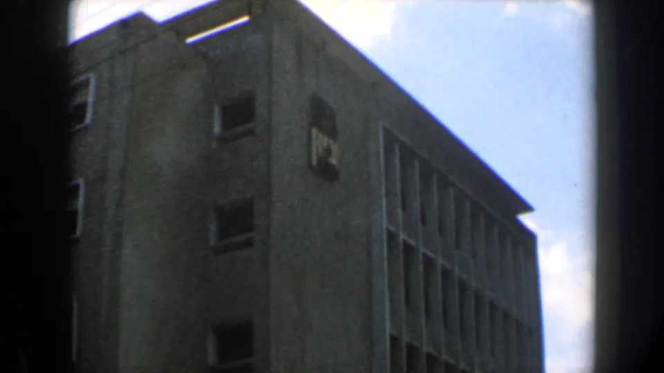 Handheld 8mm view of a concrete building corner in Israel, showing small rectangular windows and a bright blue-white sky.