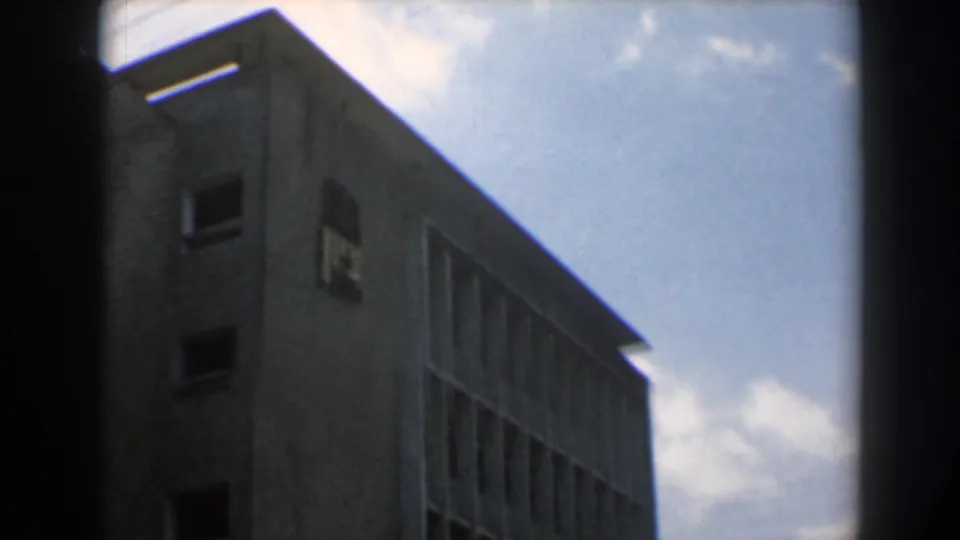1960 Israel 8mm frame with a close, low-angle look at a modern concrete facade, window grid, and cloudy blue sky.